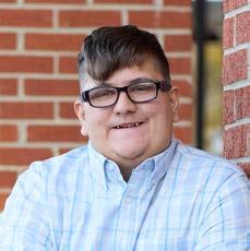 Smiling young man with short brown hair and glasses wearing a light blue plaid shirt against a brick wall.